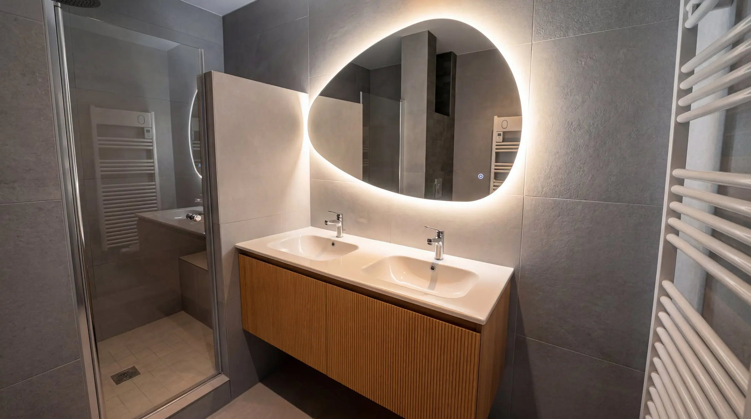 Salle de bains rénovée avec grand miroir LED organique, double vasque suspendue bois cannelé et carrelage grand format gris