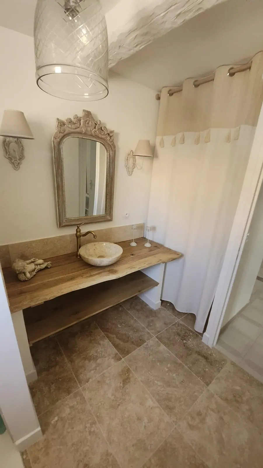 Meuble de salle de bains en bois brut à bord vivant avec vasque en pierre, robinetterie laiton et miroir ancien sculpté dans une ambiance provençale